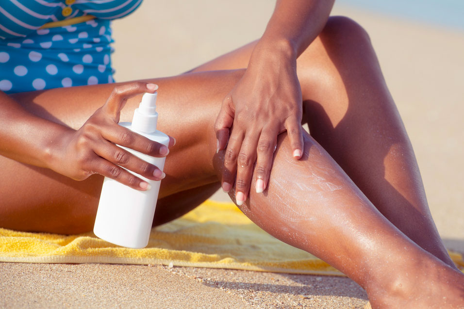 A close-up image of a woman applying sunscreen to her leg at the beach