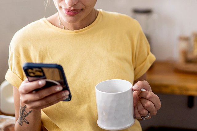 A person wearing a yellow shirt checks their phone while holding a white mug in their other hand