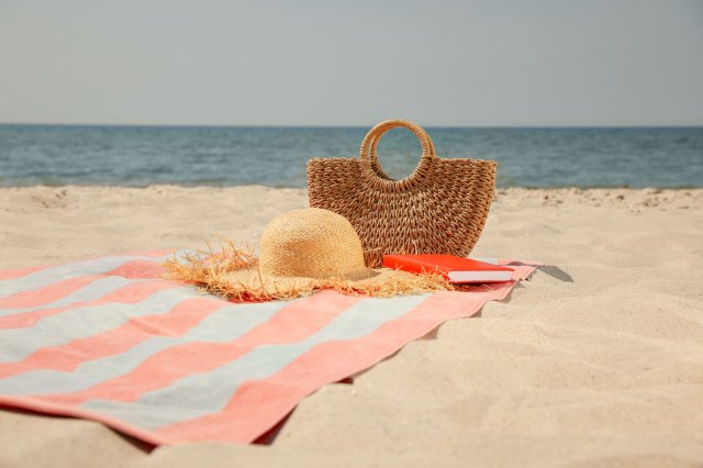 An image of a straw hat, a straw bag, and a red purse on a striped beach towel