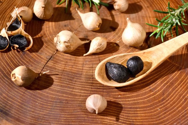 An image of black garlic cloves on a wooden surface and on a wooden spoon