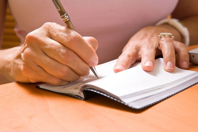 A pair of hands fills out a check in a checkbook resting on a wooden surface