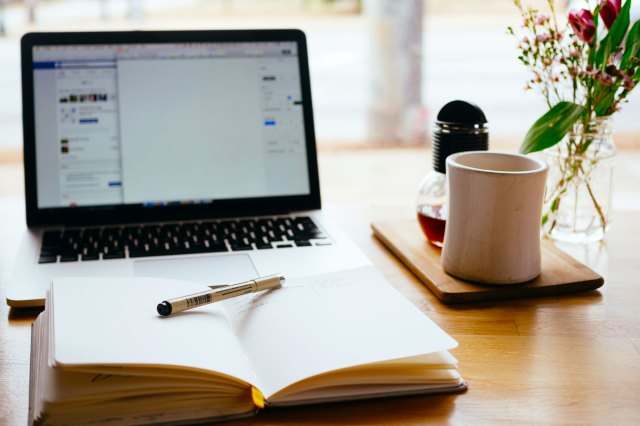 An image of a laptop, a notebook, and a white mug on a wooden table
