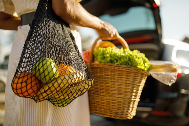 A close-up image of a woman holding a basket with vegetables with a bet bag of fruit over her shoulder
