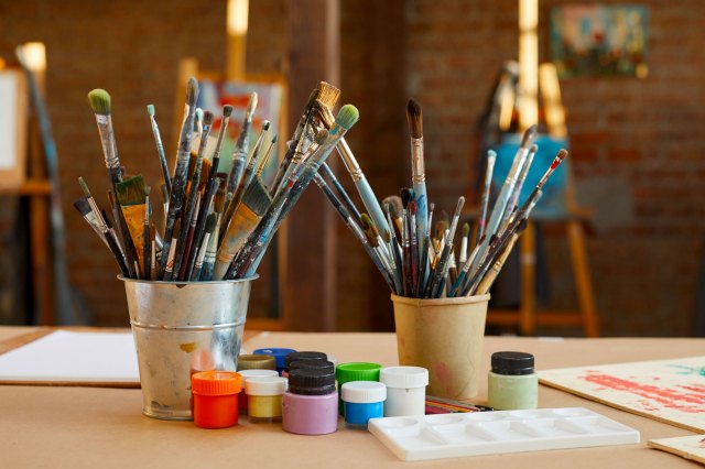 Two cups full of paintbrushes on a wooden surface with small pots of paint sitting in front of them