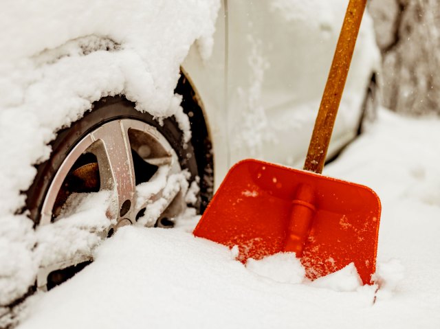 An image of a red shovel in the snow next to a snow-covered white car