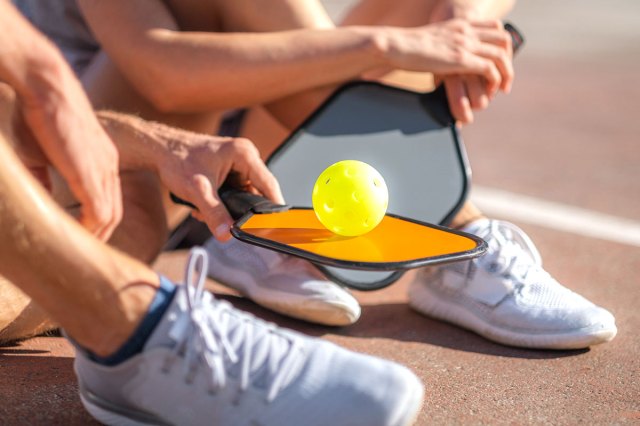 An image of two people sitting on an athletic court holding paddle ball paddles