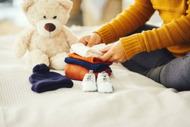 A person folds baby clothes while a teddy bear sits nearby