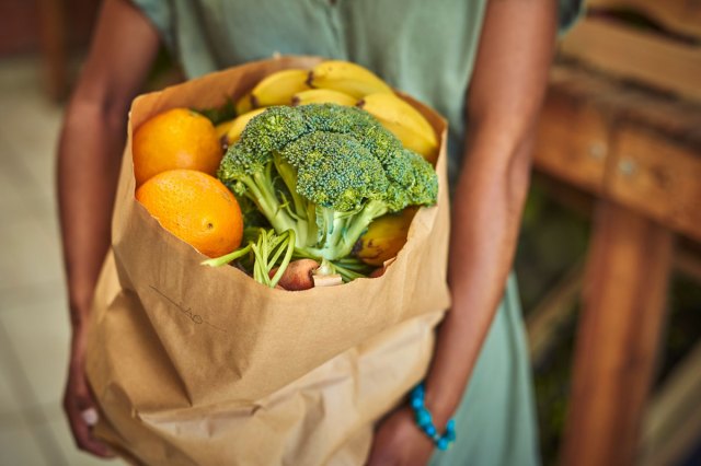 An image of a woman holding a brown shopping bag of fruits and vegetables