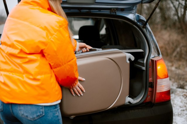 An image of a woman in an orange jacket loading a suitcase into the back of a car