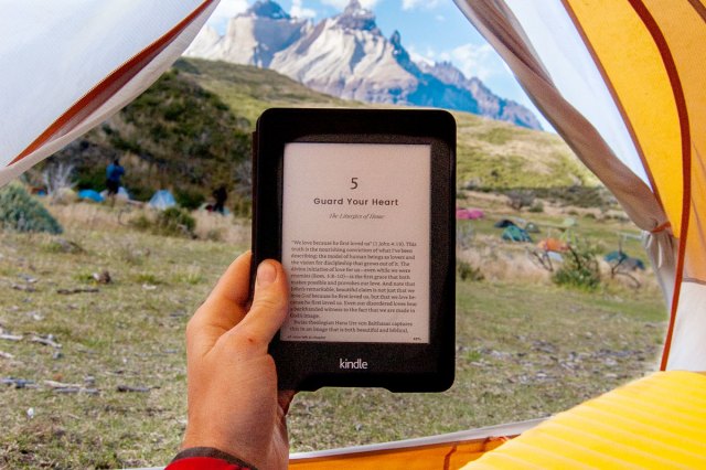 A person holds their e-reader against a natural landscape at the opening of their tent