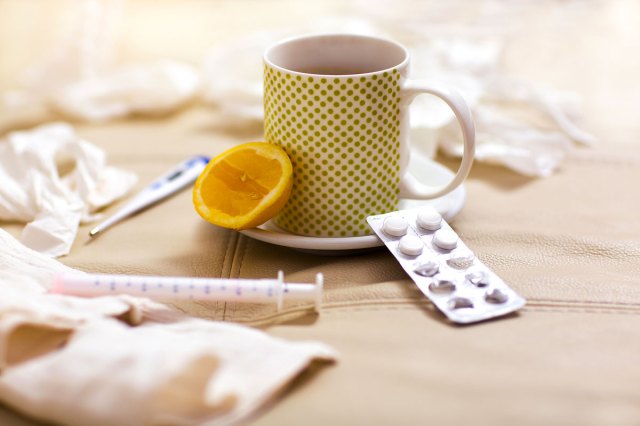 A cup of tea sits on a surface with a slice of lemon, pills, and a syringe nearby