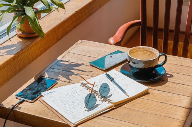 An image of a cup of coffee, a notebook with sunglasses an a pen on it, a cell phone, and a wallet on a wooden table with a chair