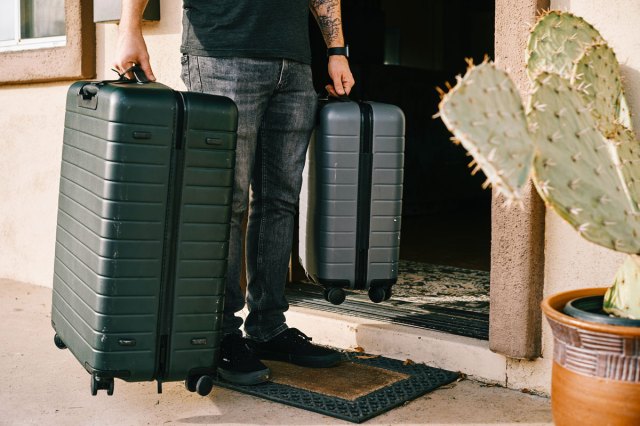 An image of a man in black denim jeans and black leather shoes standing beside black luggage bag