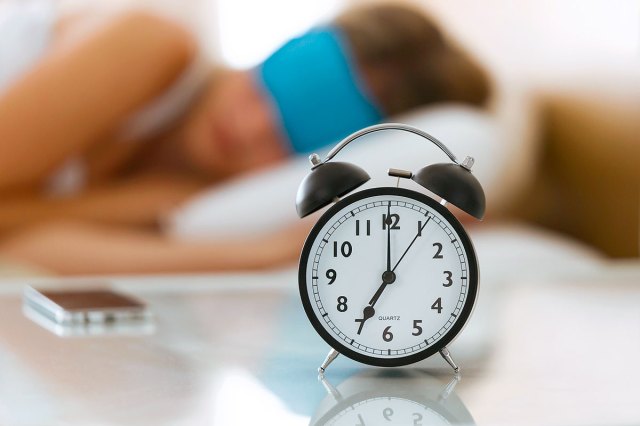 A person sleeps with an eye mask on while an alarm clock sits on a table in the foreground