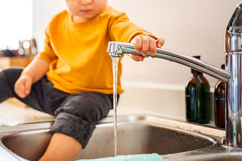 An image of a toddler sitting on the counter and holding a running kitchen faucet