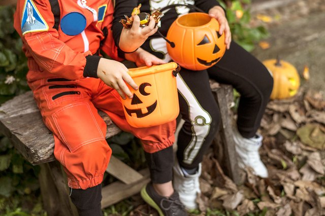 An image of two kids in Halloween costumes with jack-o'-lantern candy buckets sitting on a bench outside