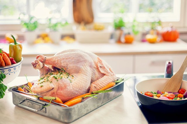 An image of a turkey in a roasting pan on the counter next to vegetables being cooked on the stove