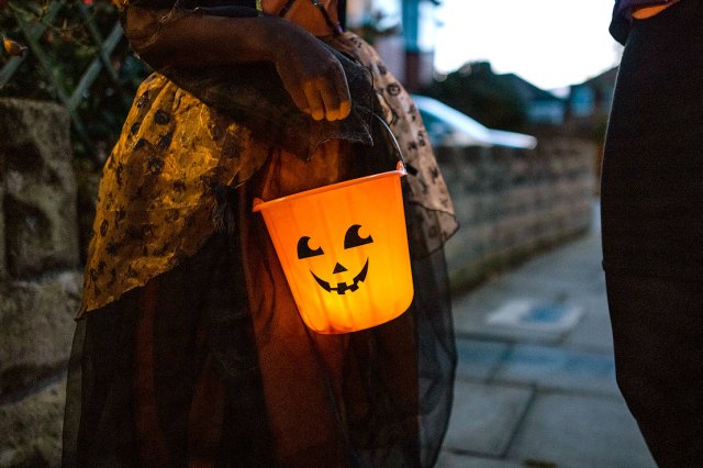 An image of a girl in a Halloween costume holding a light-up jack-o'-lantern candy bucket