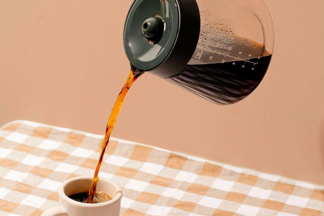 An image of a coffee from a pot being poured into a mug on a gingham table cloth