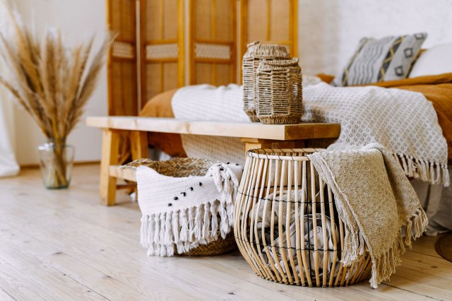 An image of a bohemian bedroom with textile sheet on bed, wooden bench seat, bamboo dressing screen, dry plants in vase, and wicker basket