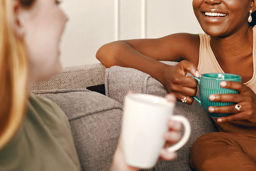 An image of two women with coffee mugs sitting on the couch