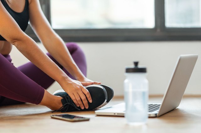 An image of a woman sitting on the floor stretching in front of a computer and water bottle