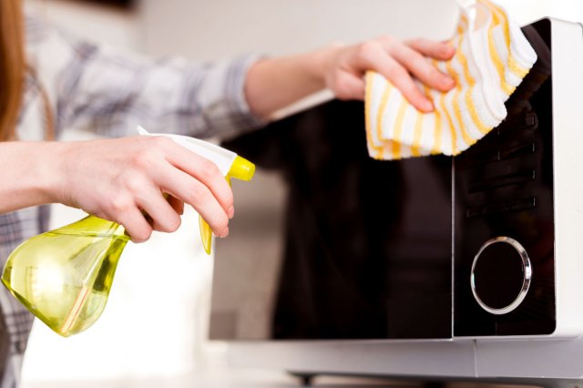 An image of a person holding a cloth and spraying cleaner on a microwave