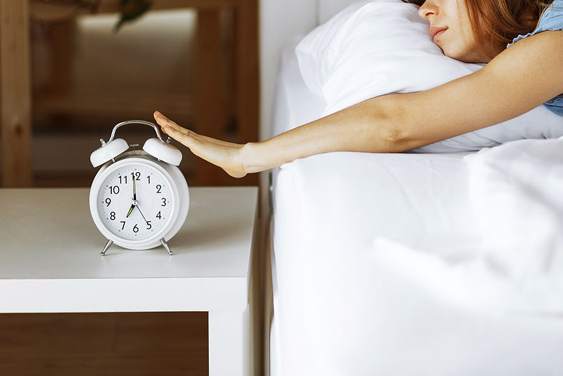 An image of a woman in bed hitting a white alarm clock
