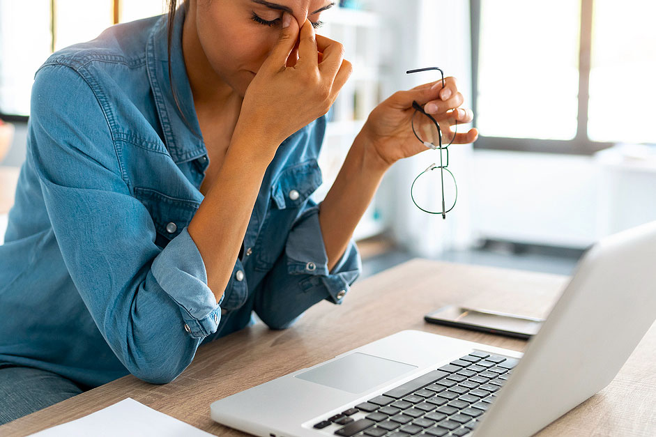 An image of a woman sitting in front of a laptop pinching the bridge of her nose in distress