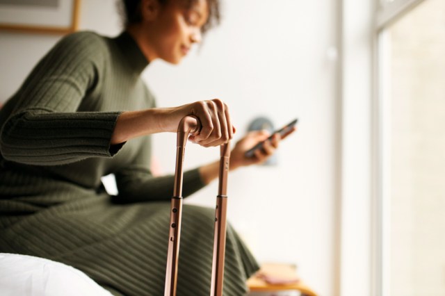 An image of a woman sitting on a bed looking at her phone and holding onto a carry-on suitcase