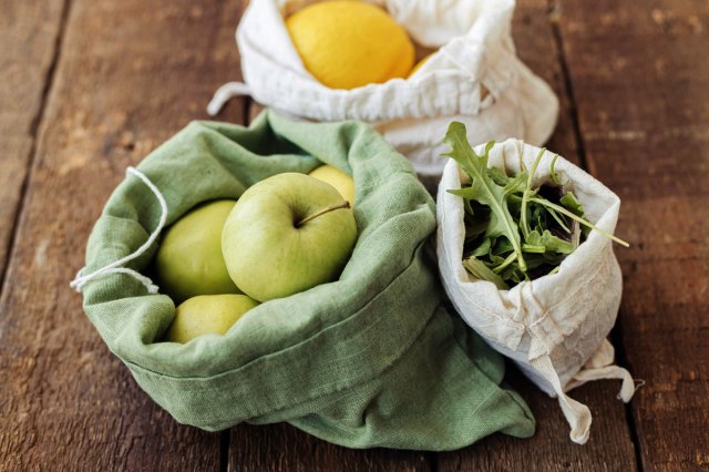 An image of a green cloth bag of apples, a white cloth bag of arugula, and a white cloth bag of lemons