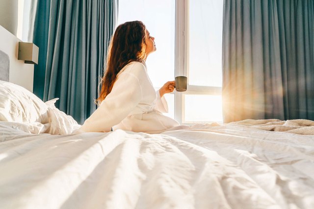 An image of a woman wearing a white robe and holding a coffee mug while sitting on a bed in the the morning