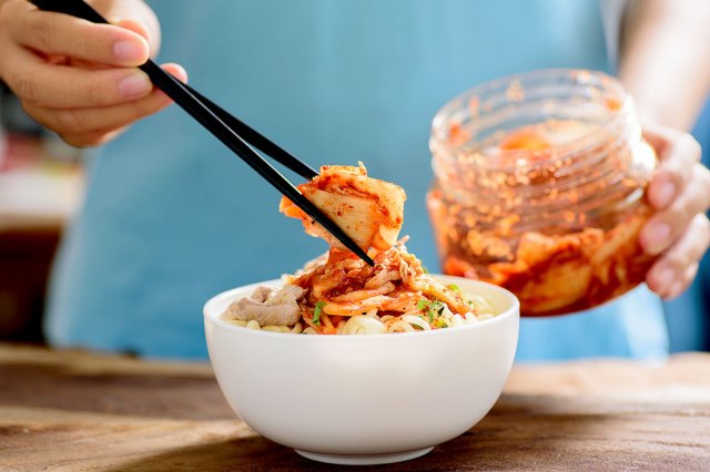 An image of a person adding kimchi with chopsticks to a bowl of ramen