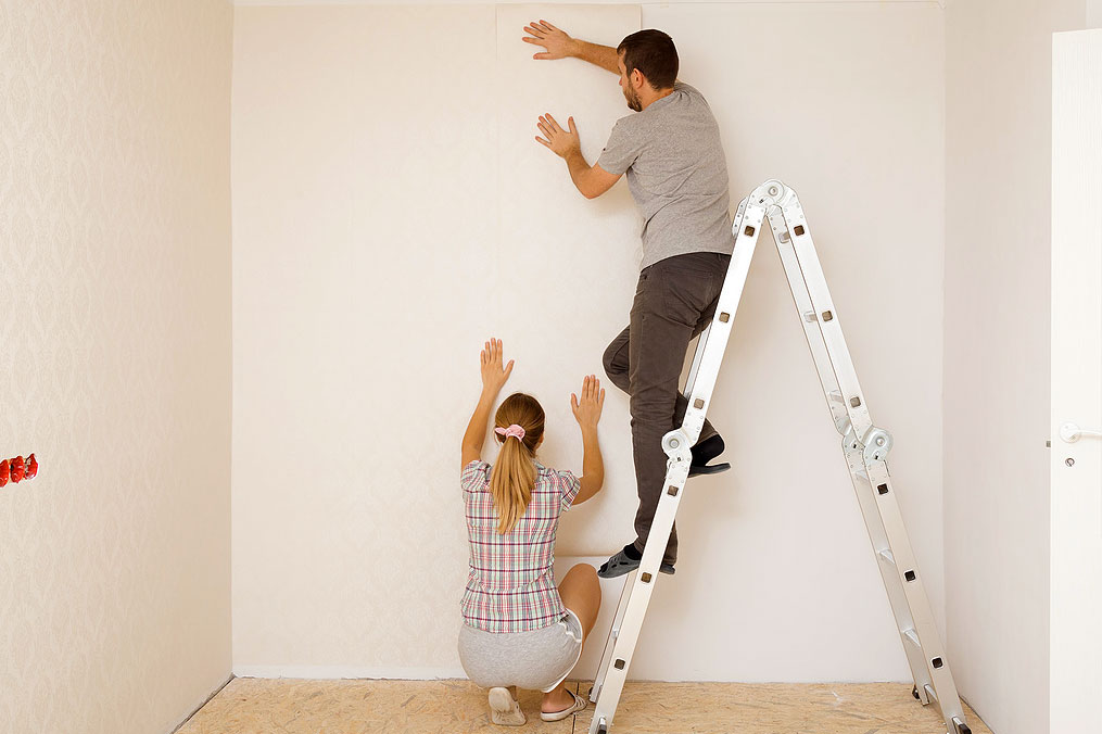 An image of a woman squatting on the floor and a man on a ladder hanging wallpaper
