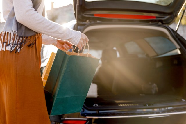 An image of a woman holding a shopping bag next to a car with an open trunk