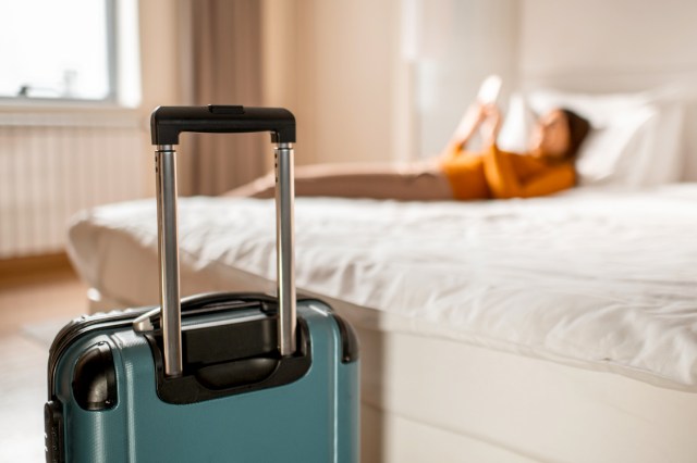 An image of a blue suitcase with a woman lying on a bed in the background