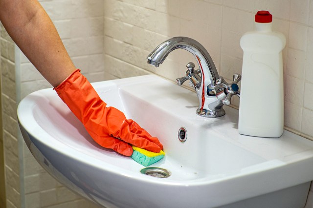 An image of a person wearing an orange rubber glove cleaning a white sink with a sponge