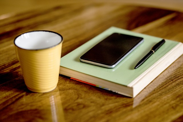 An image of a yellow coffee mug and a green book with a phone and pen resting on it