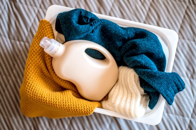 An image of sweaters and a bottle of detergent in a white laundry basket on a bed