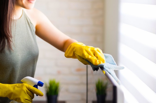 An image of a woman wearing yellow rubber gloves and cleaning blinds with a blue cloth