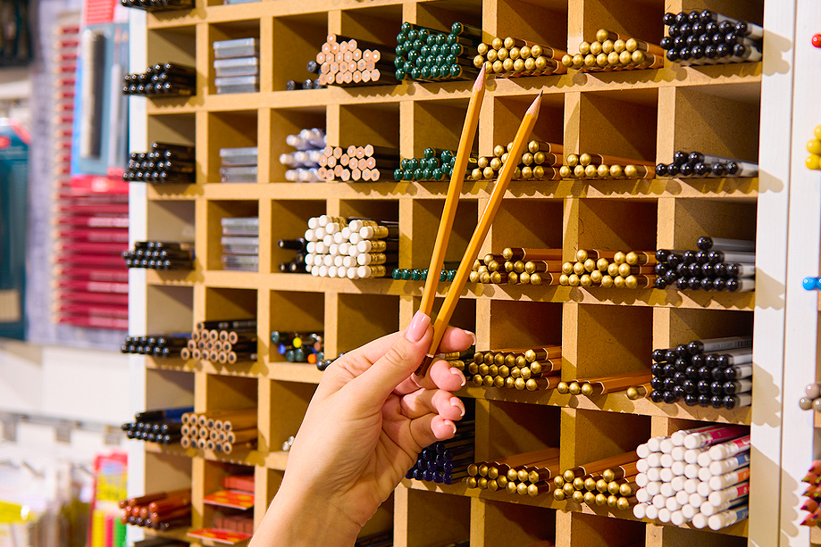 An image of a person holding two pencils in front of a shelving unit of pencils