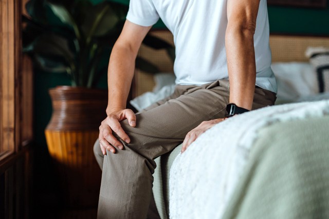 An image of a man sitting on a bed holding his knee