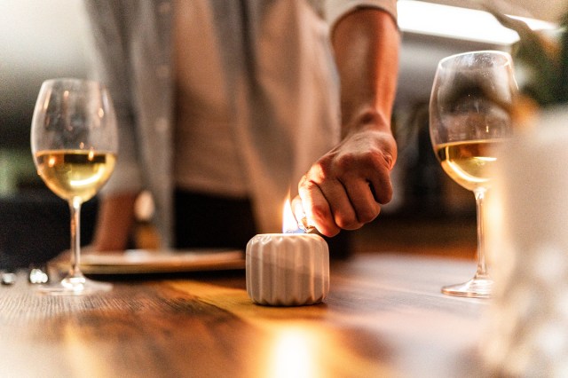An image of a person lighting a candle on a wooden table with two glasses of wine