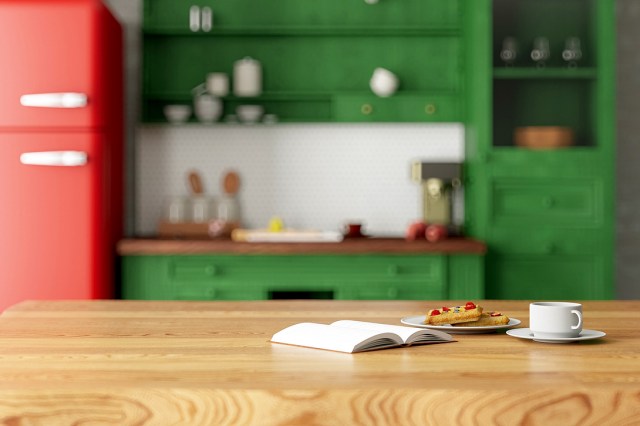 An image of a wooden counter with an open book, a plate with waffles, and a white coffee mug against a green kitchen in the background