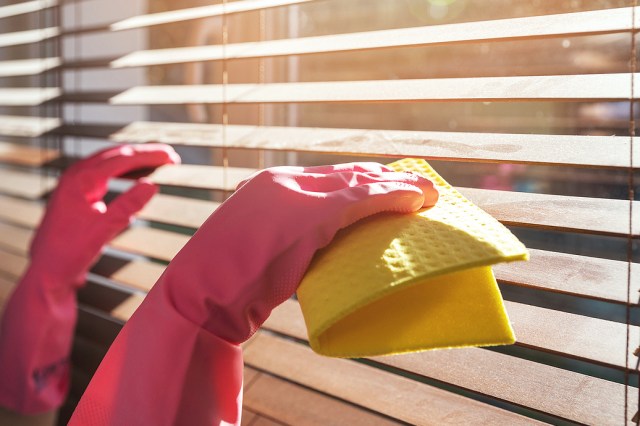 An image of hands wearing pink rubber gloves cleaning wooden blinds with a yellow towel
