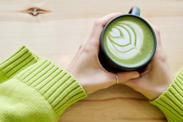 A person wearing a green sweater holds a cup of matcha