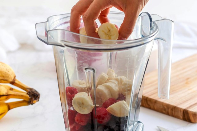 An image of a hand putting bananas in a blender with fruit