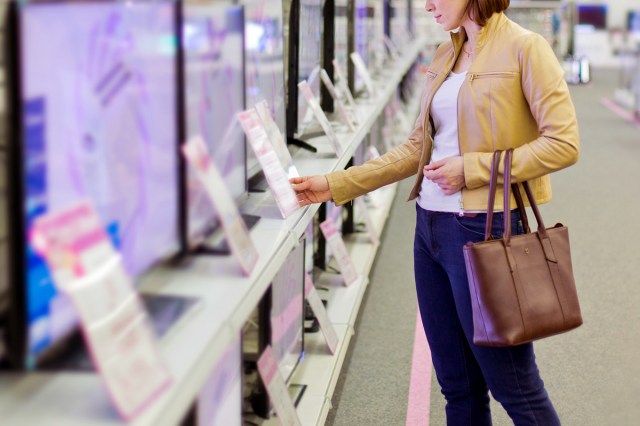 An image of a woman looking at a price sign in front of a row of televisions in a store