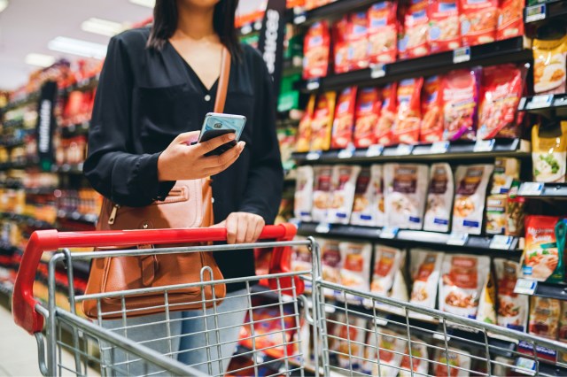 An image of a woman looking at her phone with a shopping car in a grocery store aisle