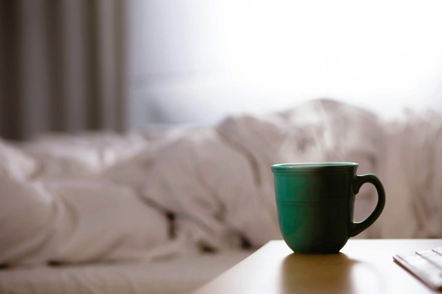 An image of a green mug on a nightstand with a bed with white sheets in the background